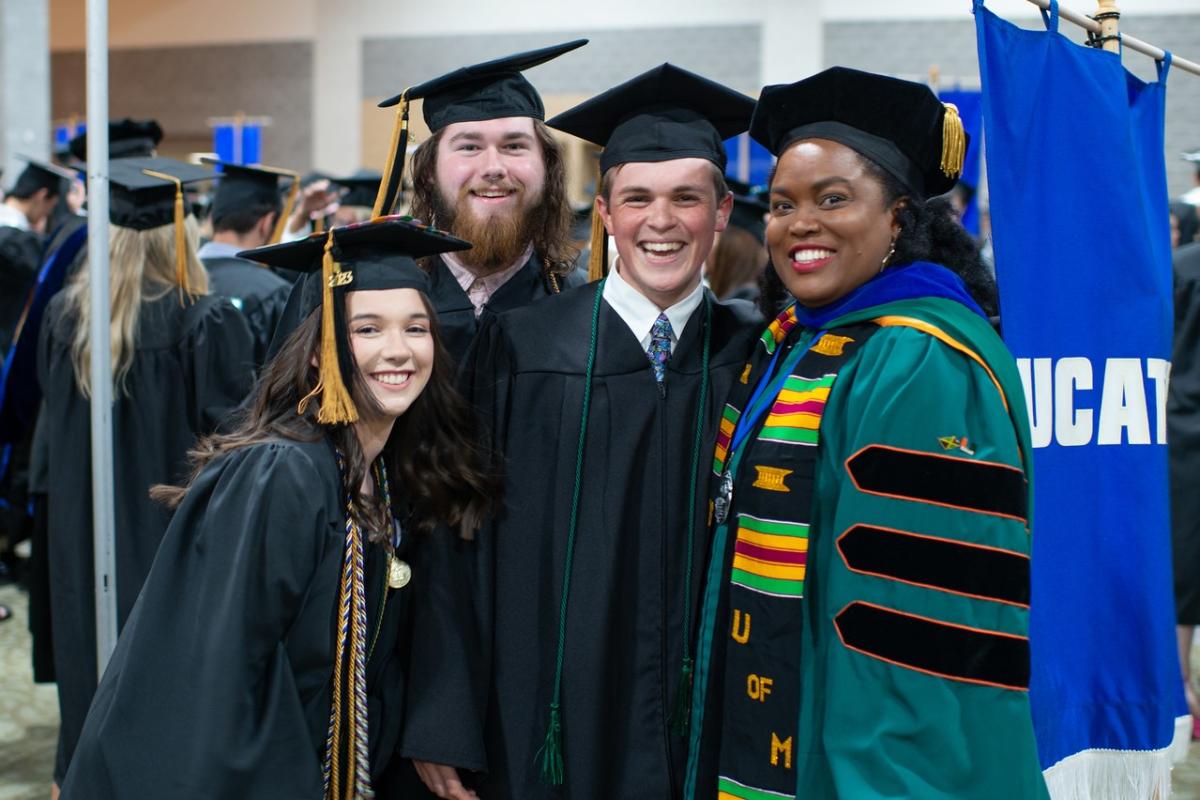 Faculty member and students smiling wearing caps and gowns at commencement.