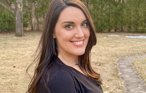 Desiree Nickel '11, a woman with long, brown hair, smiles with pine trees from an unnamed forest in the background. 