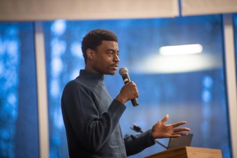 Zul Manzi, a Tanzanian-American content creator, is standing in front of a blue poster of trees. He is dressed in a long-sleeve, blue sweater and holds a microphone in his hand. He's speaking to a crowd of people who've gathered to watch his original show, "The Matimbulas".