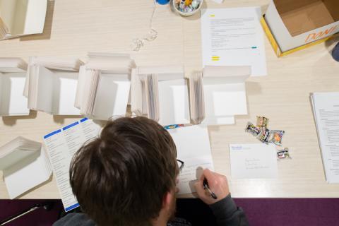 A close-up photo of a brown-haired student sitting at a white desk. He is writing hand-written thank-you cards as part of the Give a Hoot campaign. The table has stacks of cards, paper, and containers. He is writing a card, and has a pen in his hand. The angle is peeking over his shoulder with the photographer standing behind him.