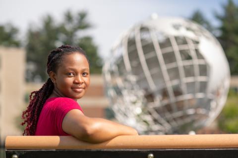 Marilyn Maison, class of 2024. She is sitting outside, on a bench in front of the silver, campus globe. The background behind her is blurred, and she has one arm slunk over the railing she leans against.