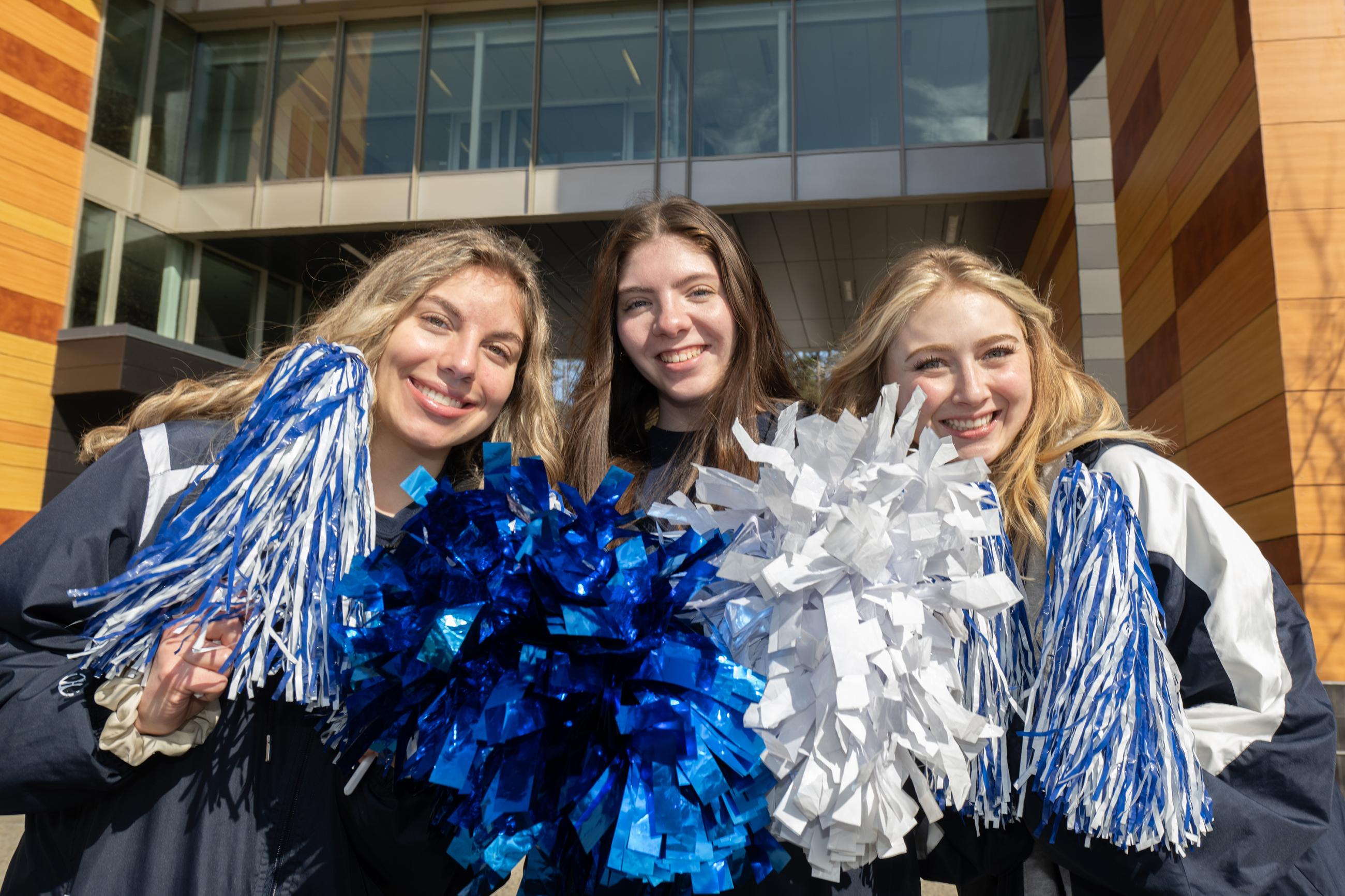 3 female WSU students, holding blue & white pom-poms, smile out in front of University Hall 