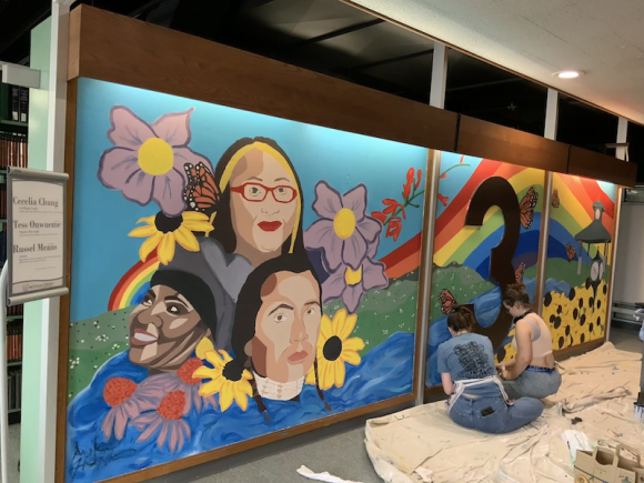 A colorful mural of sunflowers and various faces are set before a rainbow.