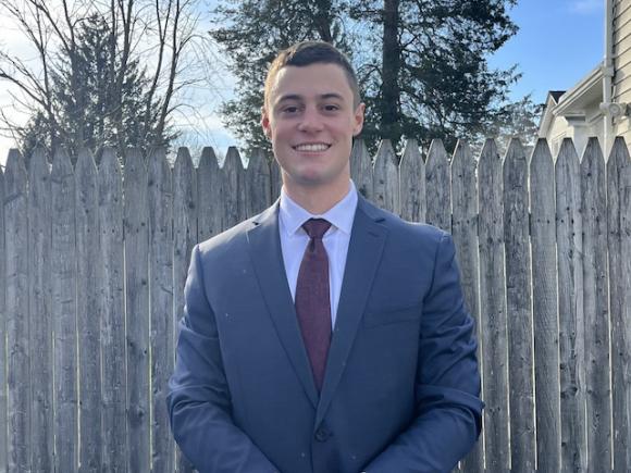 Bennett Kelly, a criminal justice major, is dressed in a blue shirt and smiles at the camera. He is outside, and a brown, wooden fence is behind him.