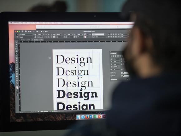 A stock photo of a student sitting in front of a computer, with him experimenting with typography in a graphic design class.