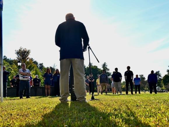 A photo from September 11's Moment of Silence Ceremony. Robert Vigneault, Assistant Director of the Office of Veteran and Military Services, is giving remarks at a small podium and microphone to a crowd of people who came to pay their respects.