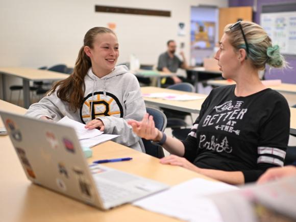 Two ABA students with laptops in a classroom talking to one another.