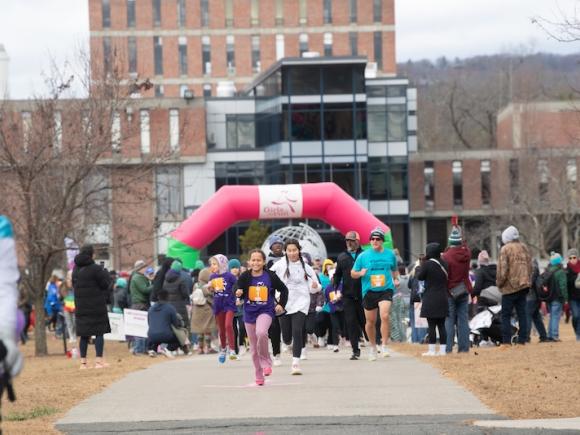 Girls on the Run, hosted on November 24. A crowd of people run towards the camera in front of the Ely Campus Center for the 5K event.