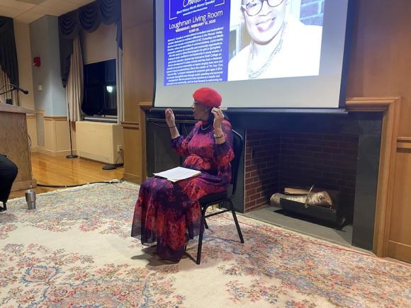 Woman sits in front of presentation screen and speaks to an audience