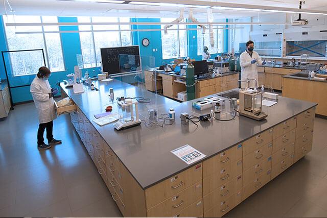 Three students wearing lab coats and protective gear work in a laboratory setting.