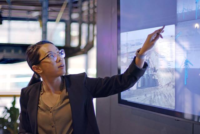 A person wearing a suit points to a large display screen while giving a presentation in a professional setting.