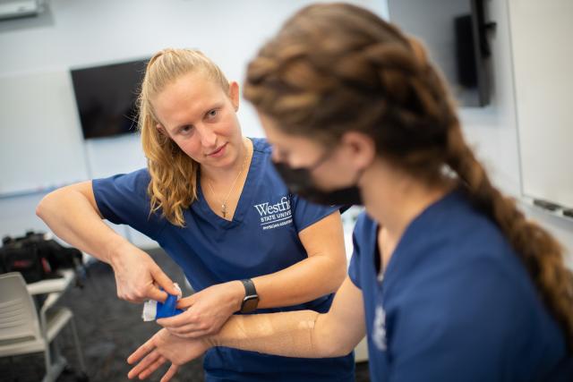 Two students in classroom splinting exercise with blue scrubs and long hair.