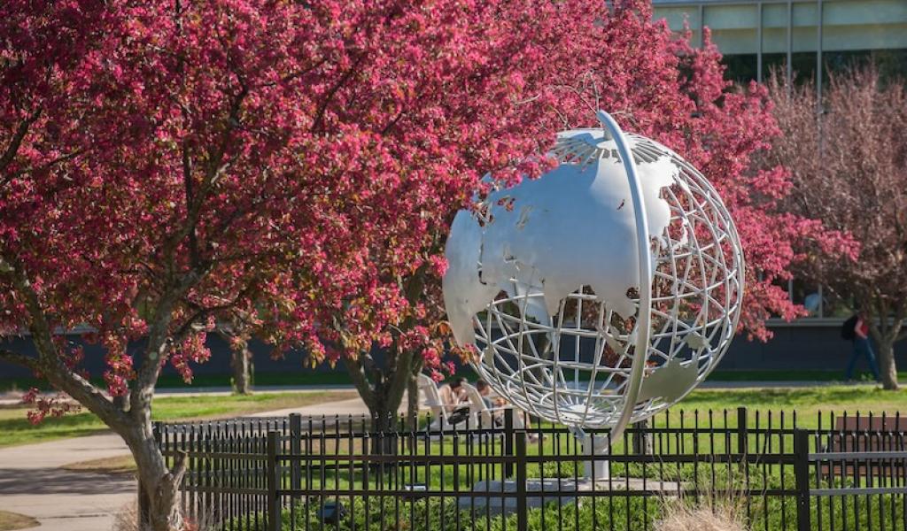 A photo of the silver, campus globe. Trees bud beside it, sprouting pink flowers. Green grass is behind the globe, and a black wrought-iron fence surrounds it.