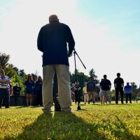 A photo from September 11's Moment of Silence Ceremony. Robert Vigneault, Assistant Director of the Office of Veteran and Military Services, is giving remarks at a small podium and microphone to a crowd of people who came to pay their respects.